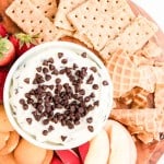 A round wooden tray holds a bowl of cream dip topped with chocolate chips, surrounded by graham crackers, waffle pieces, vanilla wafers, apple slices, and strawberries.