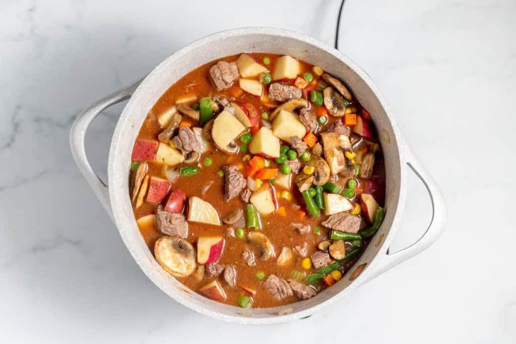 A pot filled with beef stew containing chunks of beef, potatoes, carrots, mushrooms, peas, and green beans, sitting on a light-colored countertop.