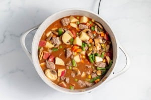A pot filled with beef stew containing chunks of beef, potatoes, carrots, mushrooms, peas, and green beans, sitting on a light-colored countertop.