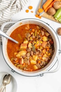 A pot of hearty vegetable and meat stew sits on a wire rack with a ladle. Ingredients like potatoes, carrots, and celery are visible. Surrounding the pot are chopped vegetables and a striped towel.