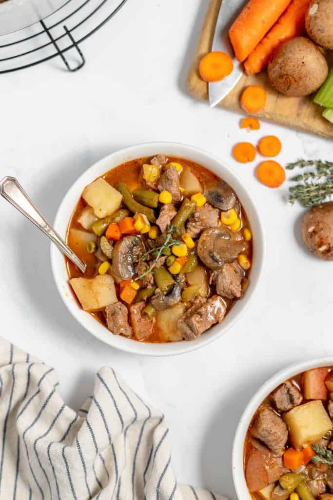 A bowl of vegetable and beef stew with potatoes, carrots, corn, green beans, and mushrooms. A spoon is placed in the bowl. Carrots, celery, and mushrooms are on a cutting board in the background.