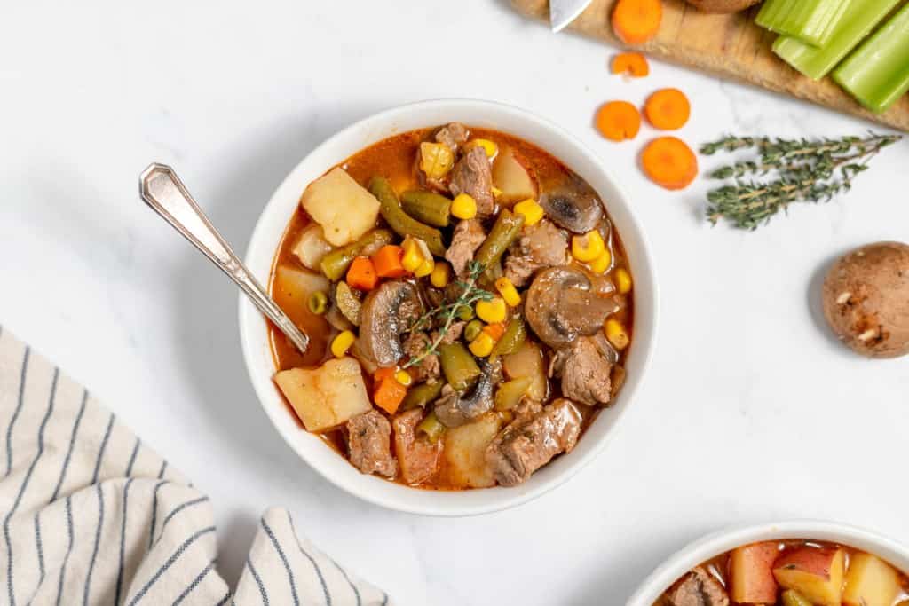 A bowl of vegetable beef stew with chunks of meat, potatoes, carrots, green beans, corn, and mushrooms. A spoon is placed in the bowl, and ingredients are seen on the table beside it.