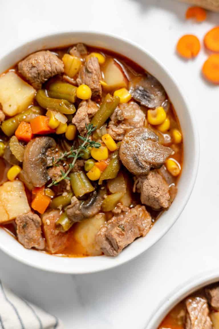 A bowl of hearty beef stew with chunks of meat, potatoes, carrots, corn, mushrooms, and other vegetables, garnished with a sprig of thyme. Sliced carrots are scattered nearby.