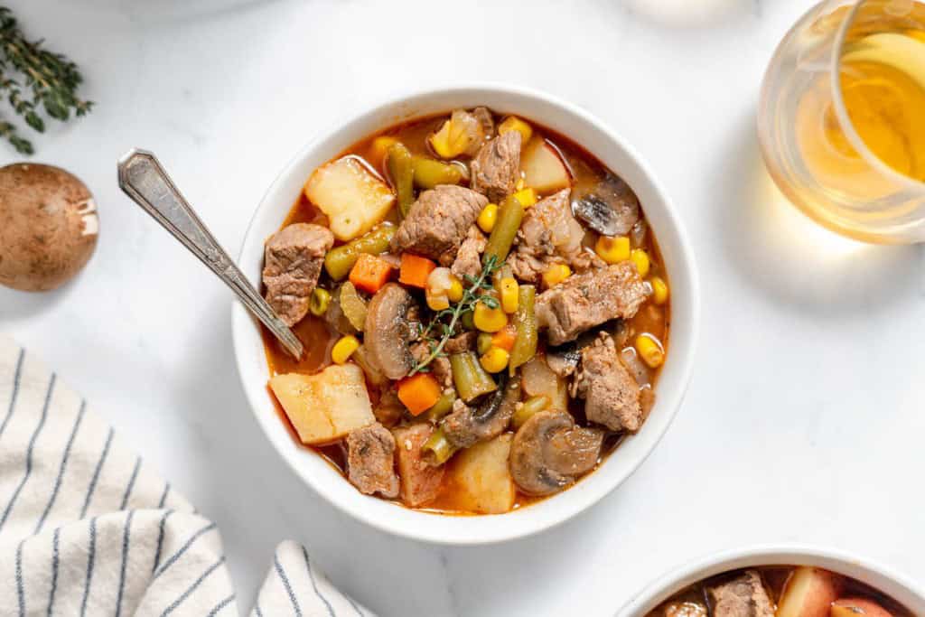 A bowl of hearty beef stew with chunks of beef, potatoes, mushrooms, corn, and other vegetables, garnished with a sprig of thyme, next to a glass of a light beverage.
