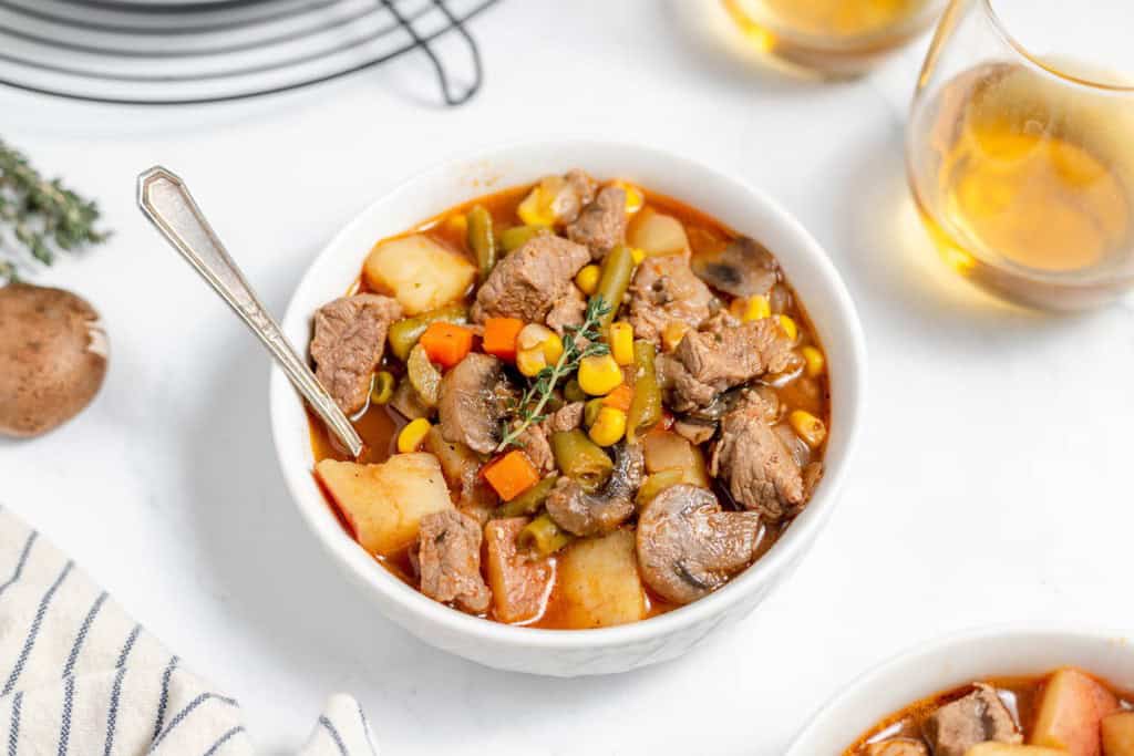 A bowl of beef stew with chunks of beef, potatoes, carrots, corn, green beans, and mushrooms, served with a spoon. Two glasses of a light beverage are in the background.