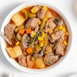 A bowl of beef stew containing chunks of beef, potatoes, carrots, green beans, corn, and mushrooms, garnished with a sprig of thyme.