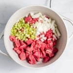 Chunks of raw beef, diced celery, and chopped onions in a white pot on a marble surface.