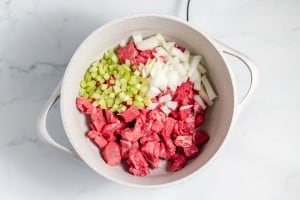 Chunks of raw beef, diced celery, and chopped onions in a white pot on a marble surface.