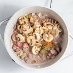 A pot containing a mixture of diced meat, mushrooms, spices, and herbs, cooking on a stovetop.