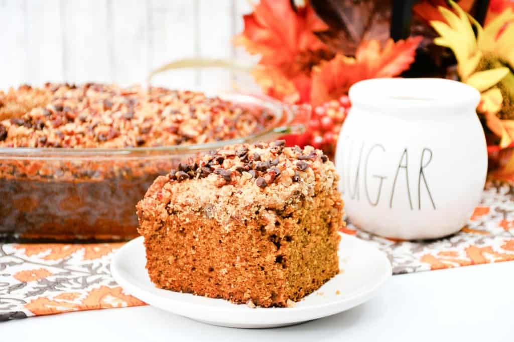 A slice of pumpkin coffee cake with crumb topping on a white plate. The remaining cake is in a glass baking dish in the background, accompanied by a sugar jar and autumn-themed decorations.