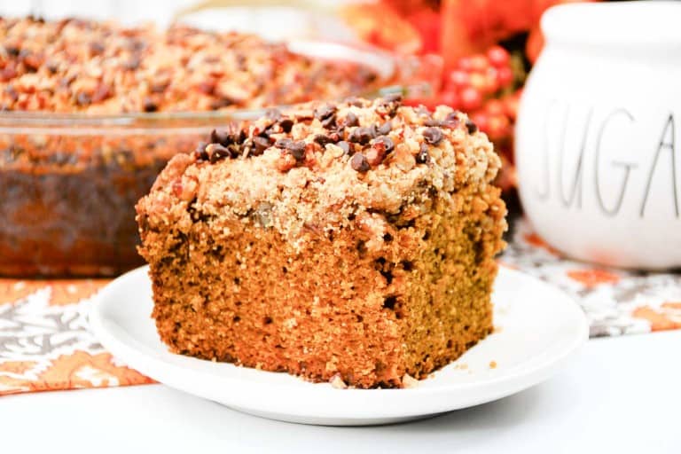 A slice of crumb-topped cake on a white plate, with a larger portion in a glass dish and a white container labeled "SUGAR" in the background.