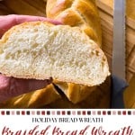 A hand holds a sliced piece of braided bread next to the unsliced braided bread wreath and a bread knife on a wooden cutting board.