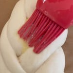 Close-up of a red brush applying egg wash to a braided dough on a baking sheet.