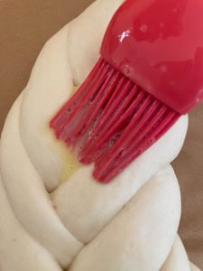 Close-up of a red brush applying egg wash to a braided dough on a baking sheet.