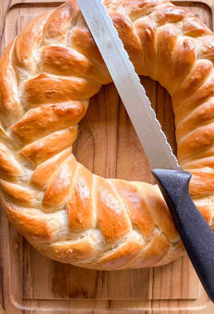 A braided circular loaf of bread on a wooden cutting board with a serrated knife resting on top.
