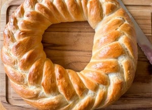 A round, braided loaf of golden-brown bread on a wooden cutting board with a serrated knife beside it.
