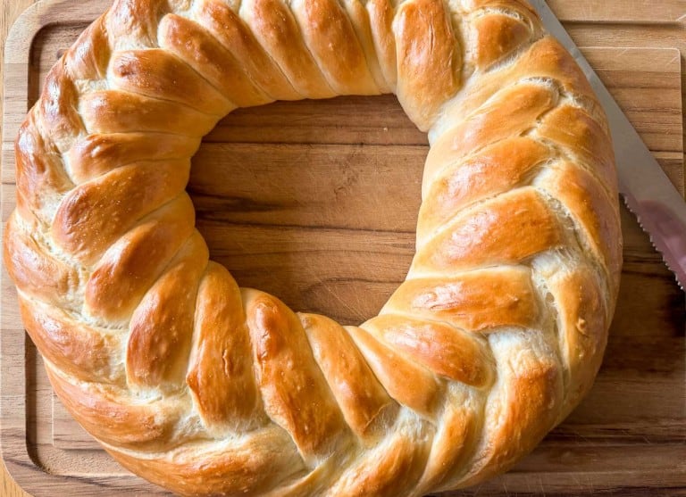 A round, braided loaf of golden-brown bread on a wooden cutting board with a serrated knife beside it.