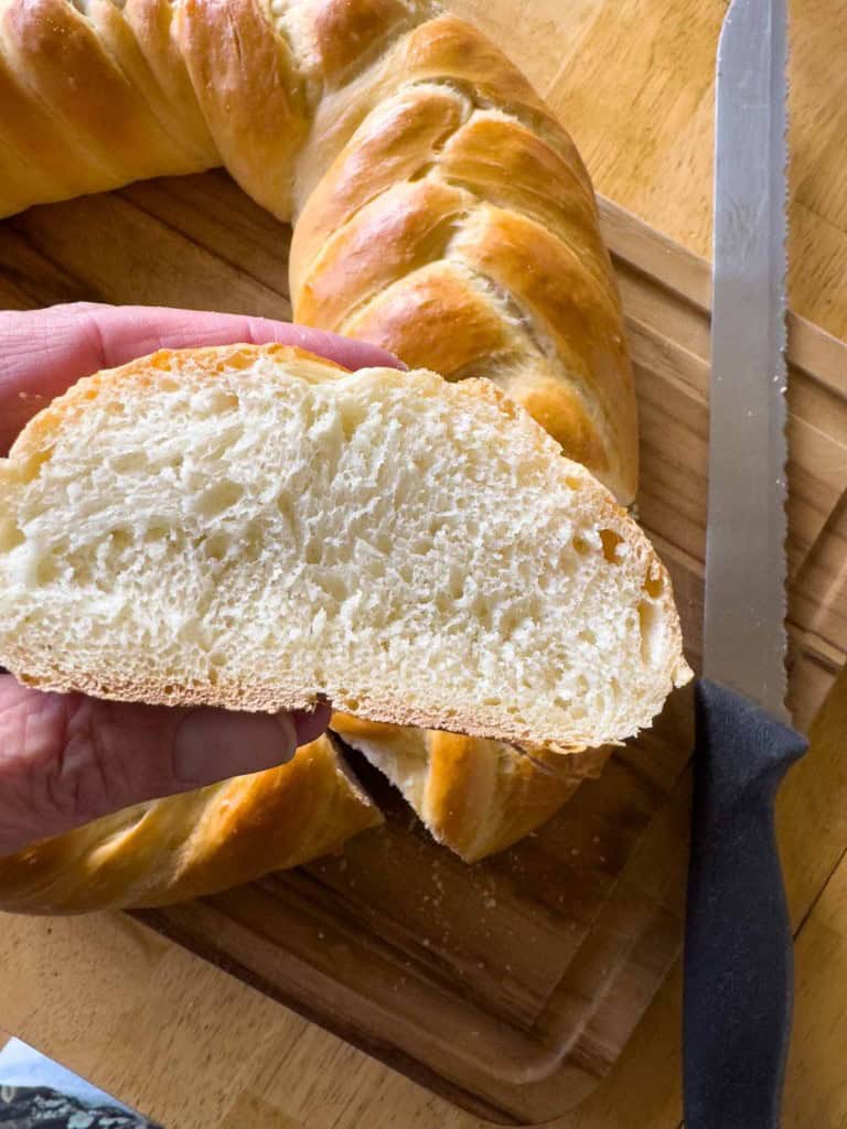 A hand holds a slice of braided bread with a soft, fluffy interior. A serrated knife rests on a wooden cutting board beside the bread.