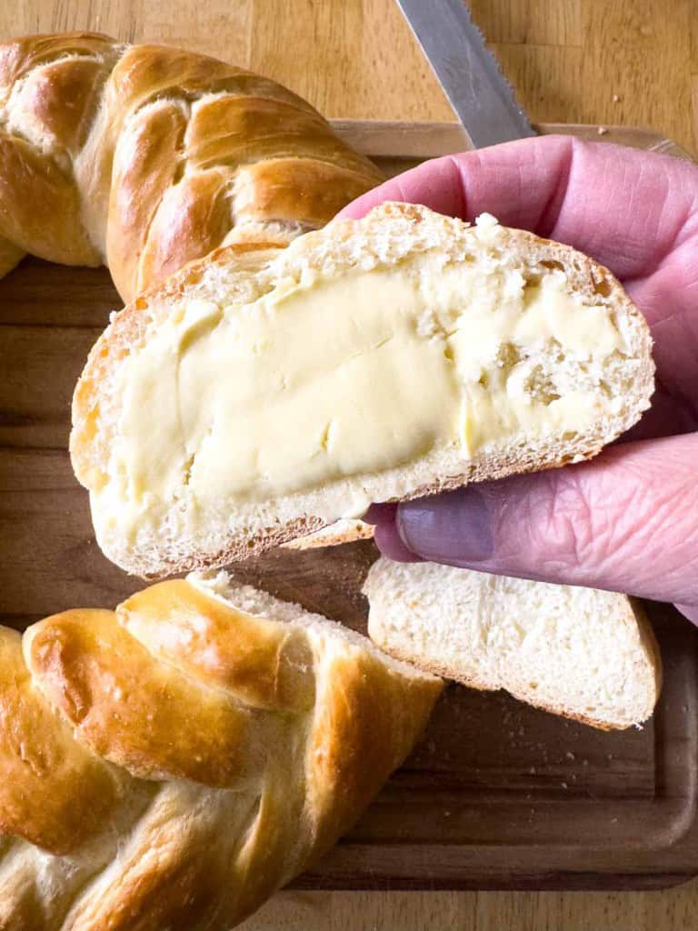 A hand holds a slice of braided bread with butter spread on it. A loaf and knife are in the background on a wooden surface.