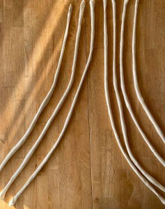 Unbaked dough rods are neatly arranged on a wooden table, illuminated by sunlight.