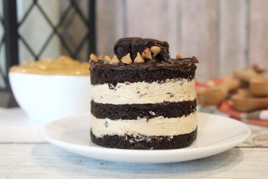 A layered chocolate cake with cream filling, topped with nuts, on a white plate. A bowl with creamy contents is in the blurred background.