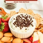 A bowl of creamy cannoli dip topped with chocolate chips is surrounded by strawberries, cookies, wafers, and apple slices on a wooden serving board.