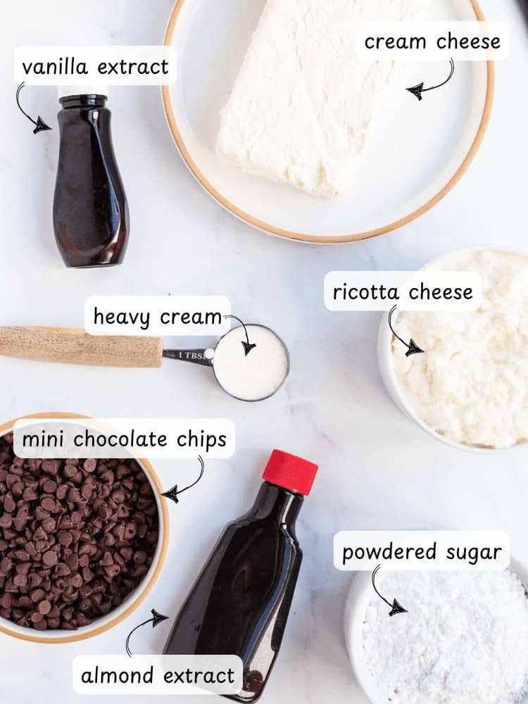 Labeled ingredients for a cannoli dip recipe: vanilla extract, cream cheese, heavy cream, ricotta cheese, mini chocolate chips, almond extract, and powdered sugar on a white surface.