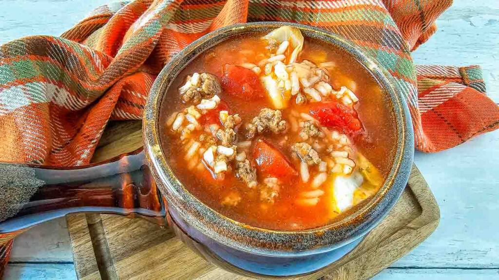 A bowl of stuffed pepper soup with ground meat, rice, and tomato pieces, infused with flavors reminiscent of classic cabbage recipes, placed on a wooden board next to a plaid cloth.