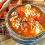 A bowl of stuffed pepper soup with ground meat, rice, and tomato pieces, infused with flavors reminiscent of classic cabbage recipes, placed on a wooden board next to a plaid cloth.
