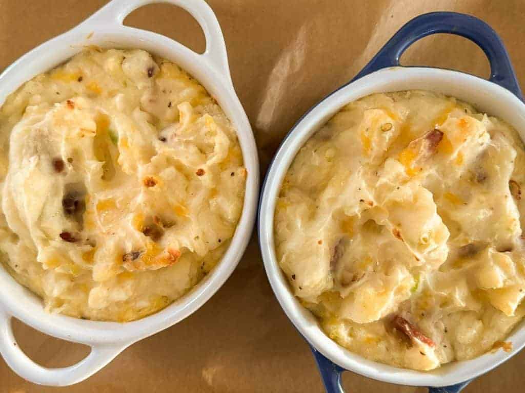 Two ramekins filled with baked mashed potatoes mixed with cheese and bacon bits on a brown surface.