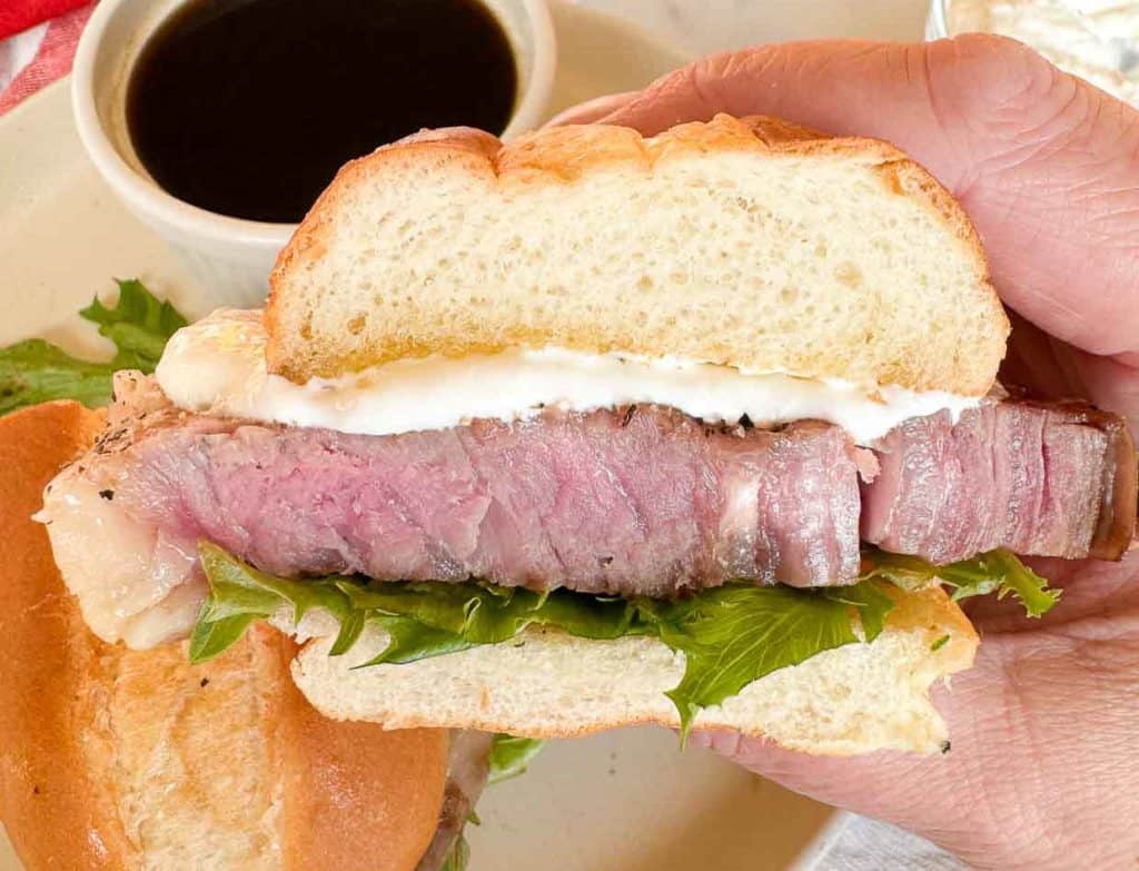 A hand holds a sandwich filled with steak, lettuce, and a white sauce. A cup of dark dipping sauce is in the background.