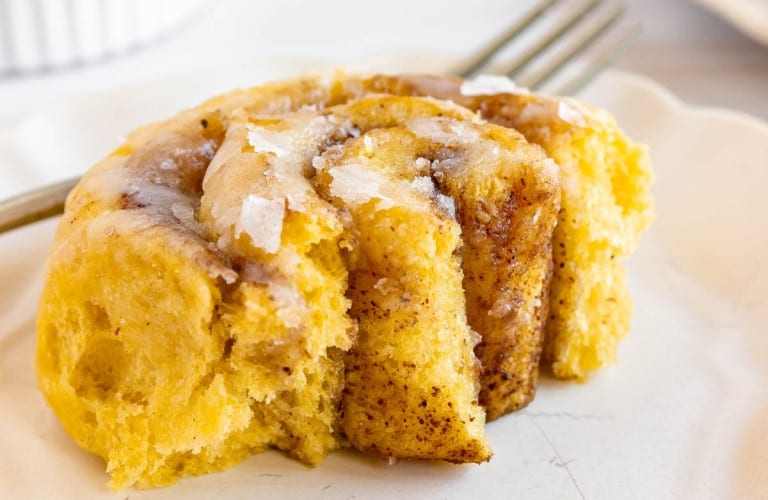A close-up of a partially eaten cinnamon roll with icing on a white plate, reminiscent of traditional Halloween treats. A fork rests beside it.