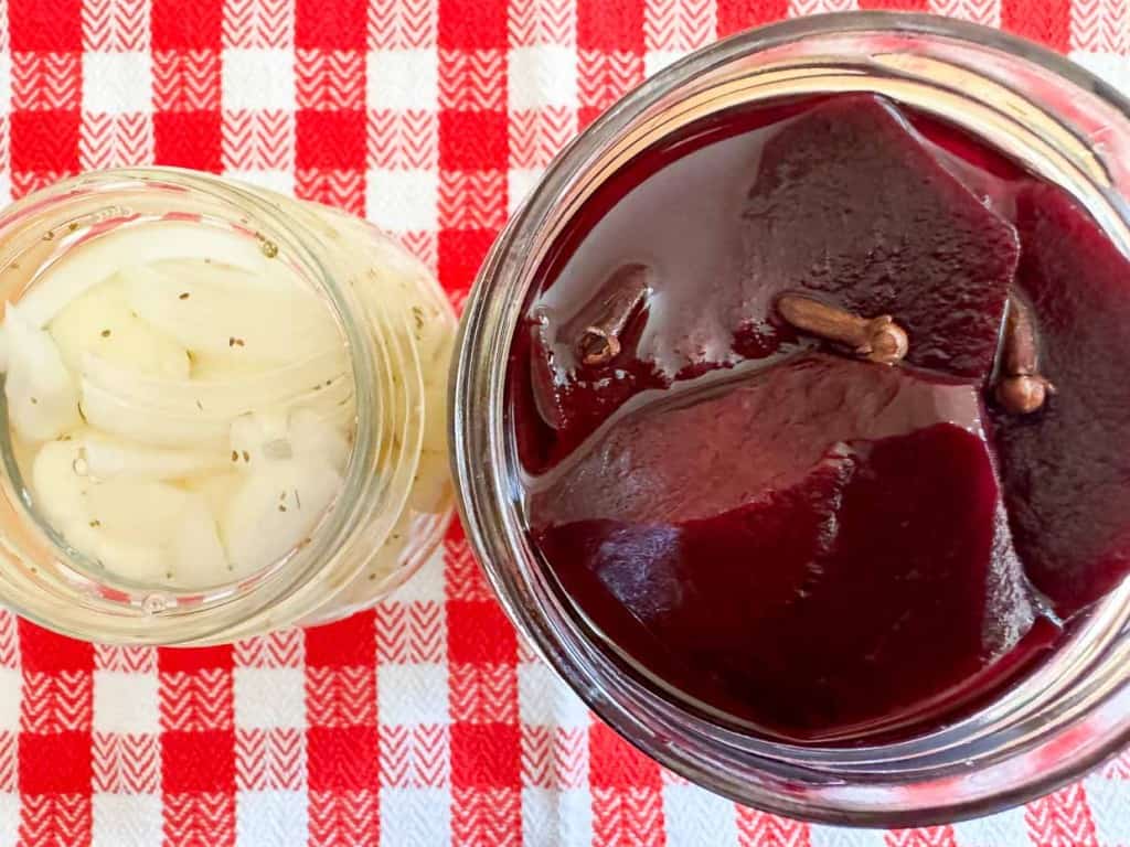 Two open jars placed on a red and white checkered cloth. The left jar contains pickled onions, and the right jar contains slices of pickled beetroot with cloves.