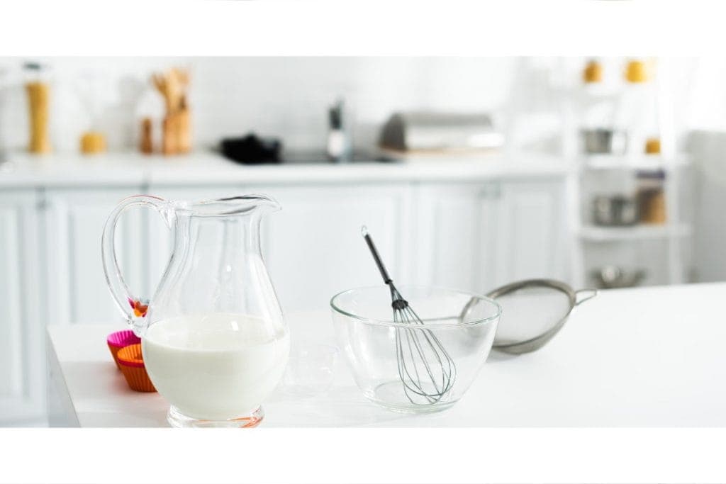 A glass pitcher of milk, a glass bowl with a whisk, and a sieve on a kitchen counter.