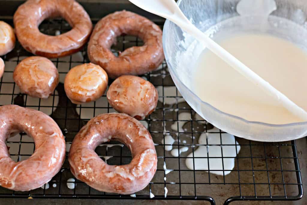 Glazed doughnuts and delightful donut holes cool on a wire rack beside a bowl of white icing with a spoon.