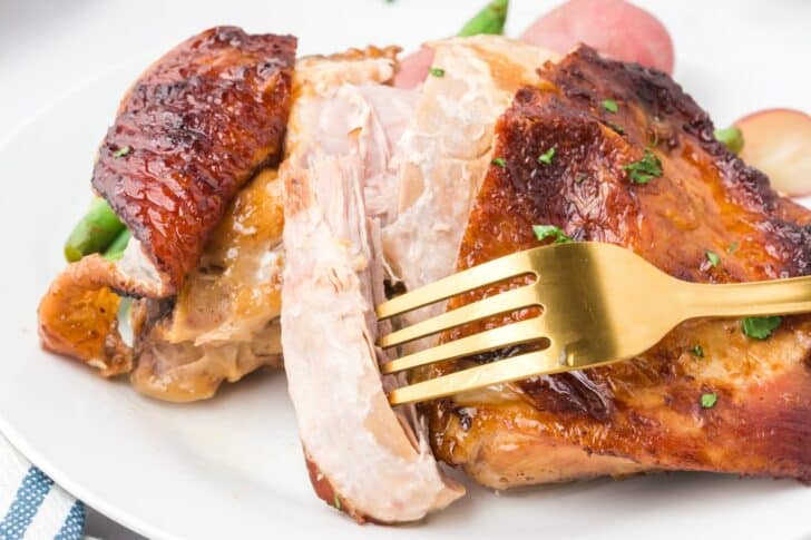 A golden fork pierces a slice of roasted turkey breast with crispy skin on a white plate, garnished with parsley—a delicious example of gourmet turkey recipes.