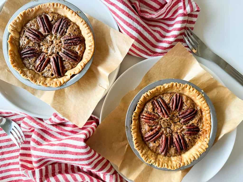 Two pecan pies rest on parchment paper, accompanied by red and white striped napkins and silver forks, capturing the essence of Thanksgiving delight.
