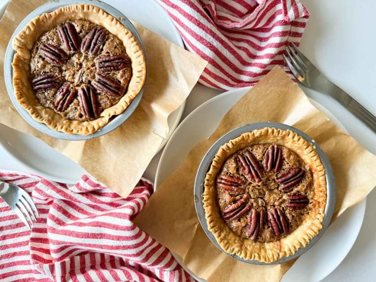 Two pecan pies rest on parchment paper, accompanied by red and white striped napkins and silver forks, capturing the essence of Thanksgiving delight.