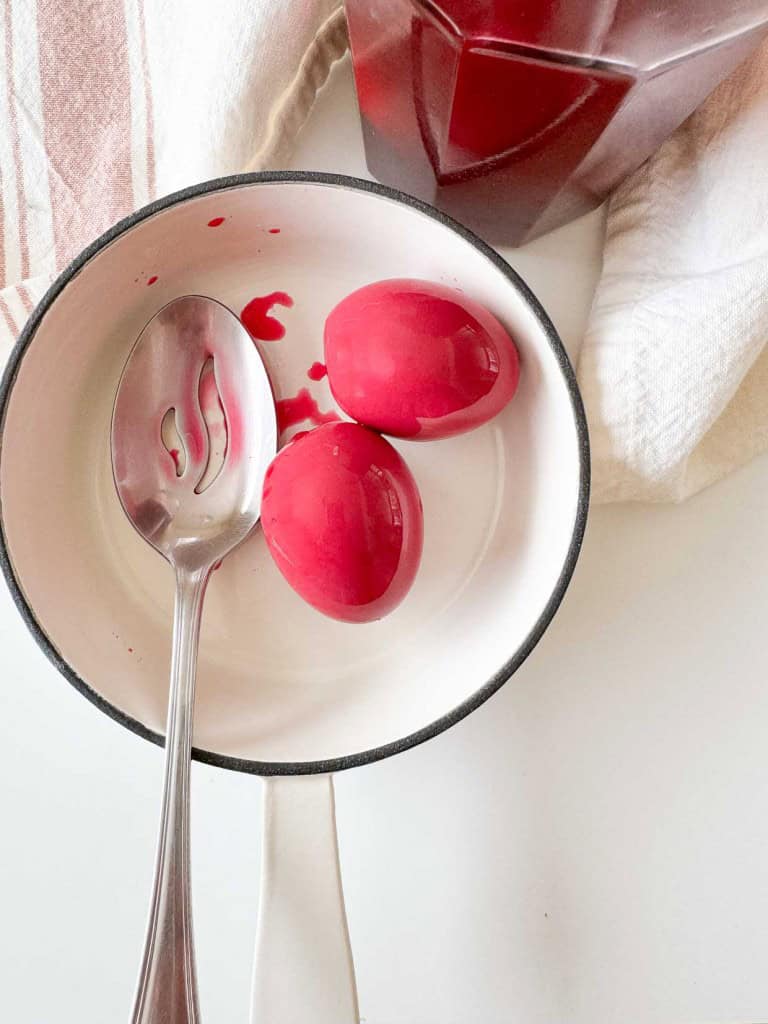 Two red-dyed eggs in a white pot with a silver spoon, nestled beside a white cloth and a red container—perfect inspiration for your next pickle recipe adventure.