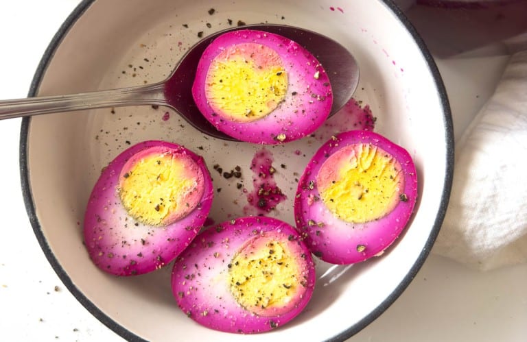 A bowl containing sliced purple pickled eggs with a spoon, seasoned with black pepper, offers a delightful twist to savory egg recipes.