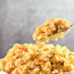 A bowl of cornbread casserole with corn, topped with a golden crust. A spoon holds a portion above the bowl.
