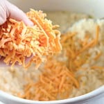 A hand adds shredded orange cheese to a white mixing bowl filled with a creamy mixture, using a wooden spoon.