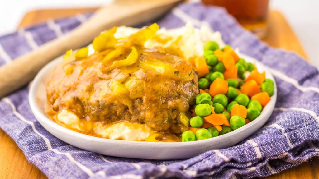 A plate with Salisbury steak topped with gravy and onions, mashed potatoes, carrots, and peas, set on a blue checkered cloth.