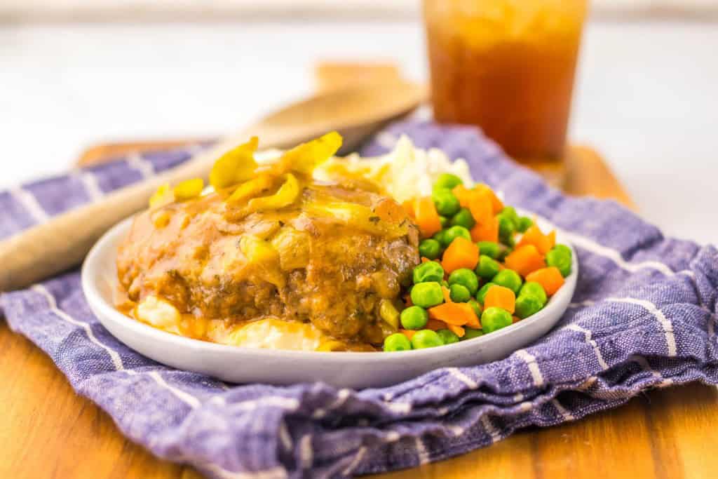 Plate of meatloaf with mashed potatoes, peas, and carrots on a blue cloth. A wooden spoon and drink are in the background.