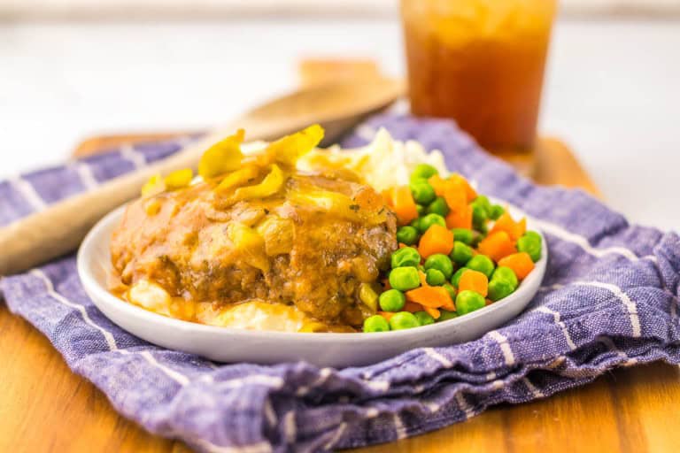 Plate with mashed potatoes, meatloaf topped with gravy and yellow peppers, alongside peas and carrots, on a blue cloth, wooden board, and iced drink in the background.