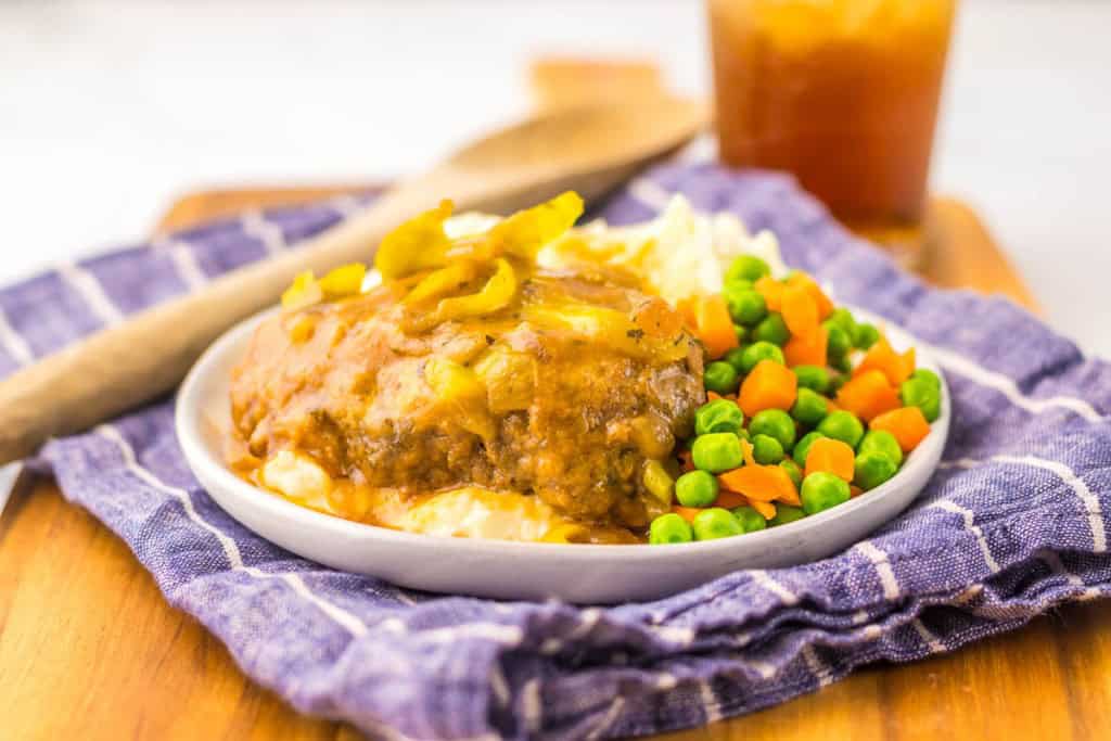 Plate of meatloaf with mashed potatoes, peas, and carrots on a blue cloth. A wooden spoon and drink are in the background.