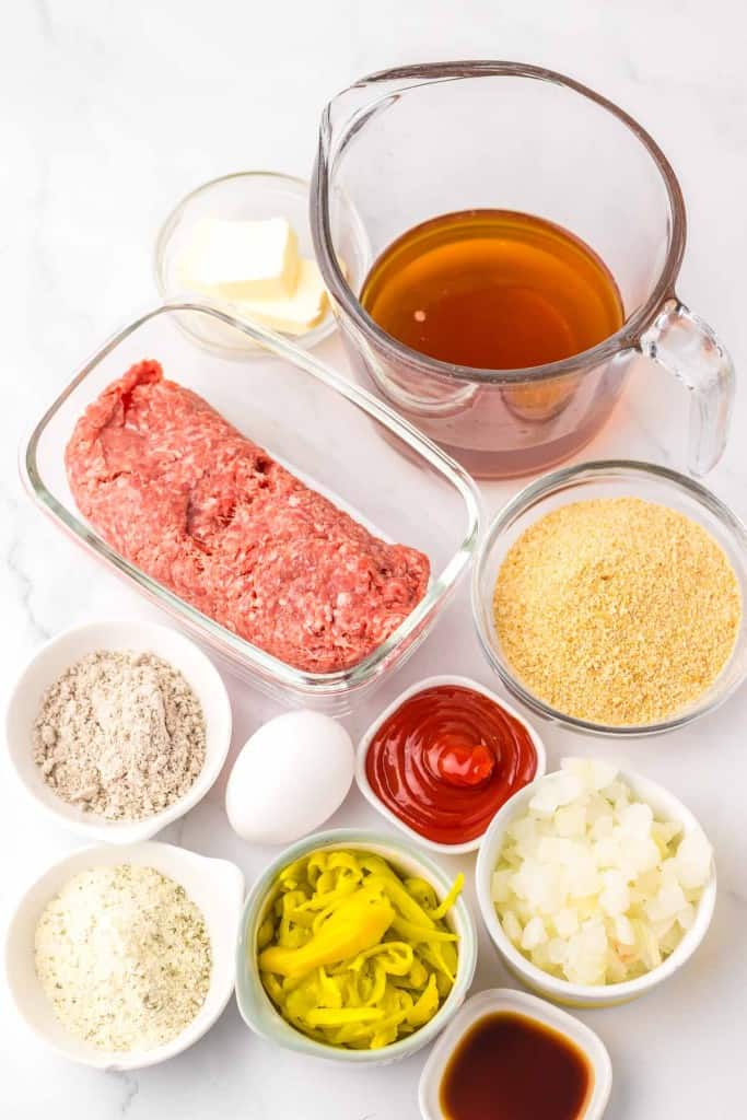 Ingredients for a recipe include ground beef, broth, breadcrumbs, flour, egg, ketchup, chopped onions, sliced peppers, butter, a seasoning mix, and sauce in small bowls and dishes.