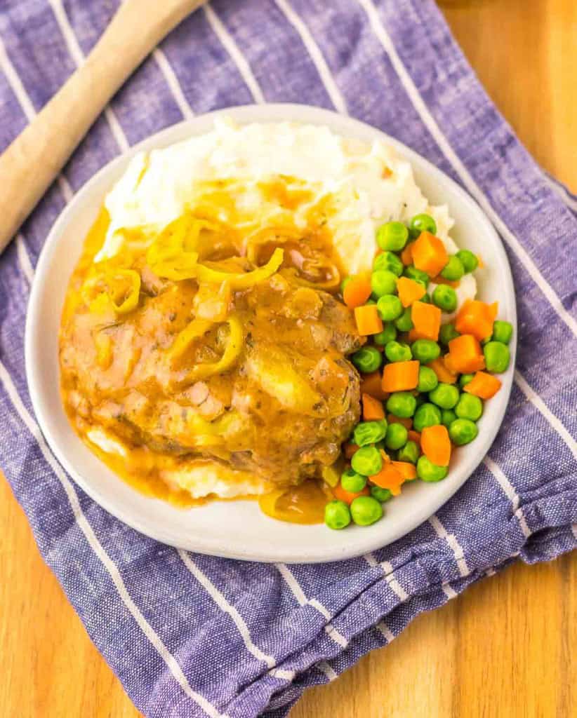 A plate with Salisbury steak and gravy, mashed potatoes, and mixed vegetables, placed on a blue-striped cloth.