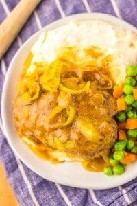 Plate with a meatloaf topped with onions and gravy, served with mashed potatoes, peas, and carrots on a blue striped cloth.