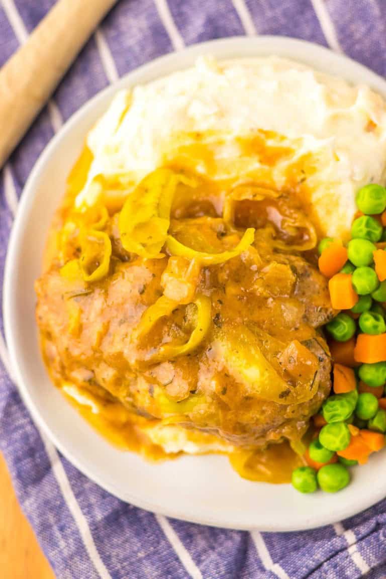 Plate with a meatloaf topped with onions and gravy, served with mashed potatoes, peas, and carrots on a blue striped cloth.